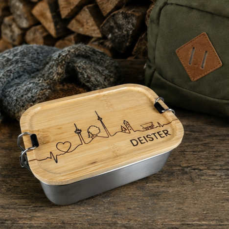 Brotdose aus Edelstahl mit Bambusdeckel und gravierter Deister-Skyline. Die Lunchbox im Format 16 x 11 cm steht auf einer Holzoberfläche und eignet sich ideal für Schule, Büro, Wandern und Outdoor-Aktivitäten in der Region Hannover.