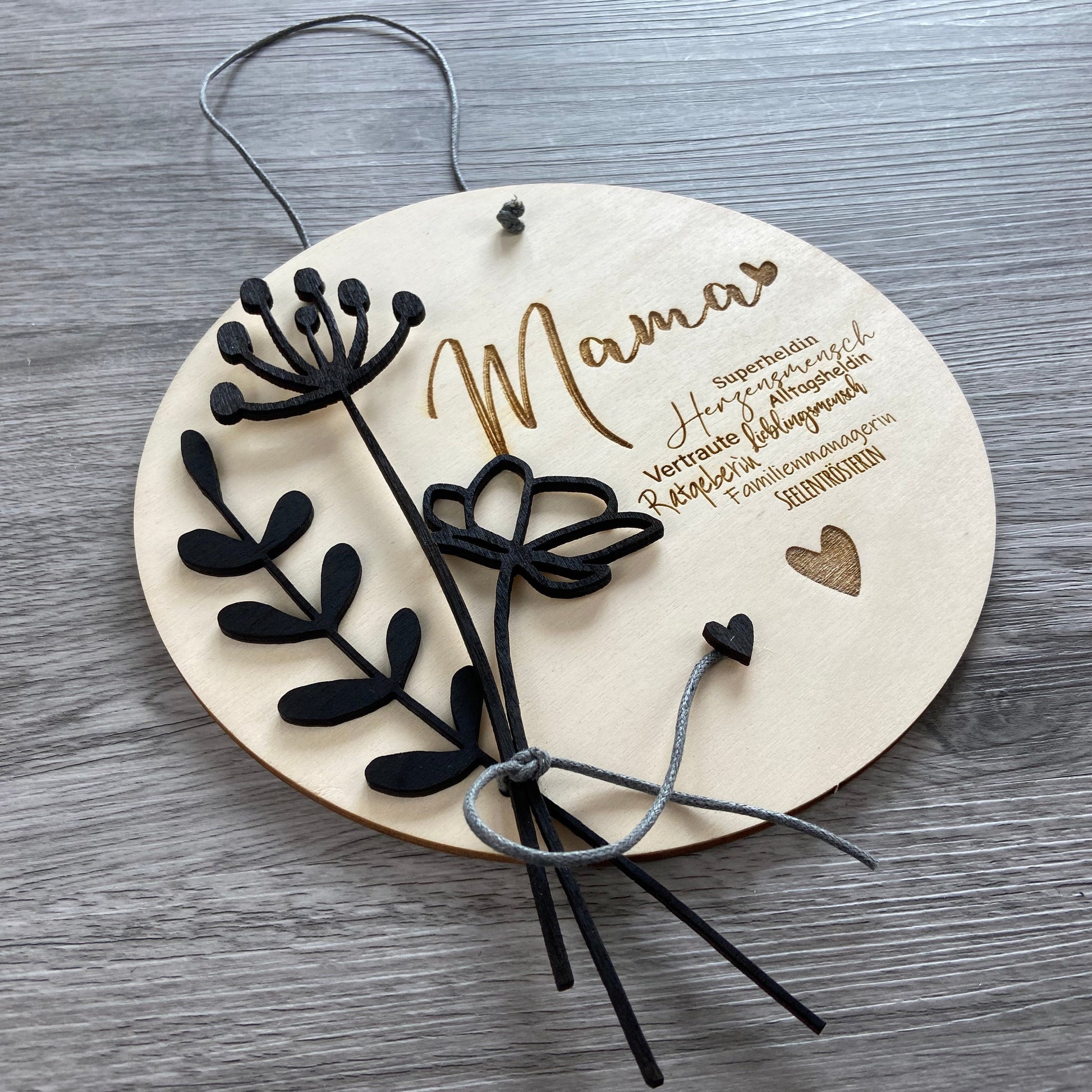 Holzschild Mama mit schwarzen Holzblumen verziert, mit Synonymen passend zu "Mama"  u.a. Herzensmensch, Superheldin, Familienmanagerin - Geschenkidee zum Muttertag mit individueller Gravur Wunschtext, leichte Schrägansicht